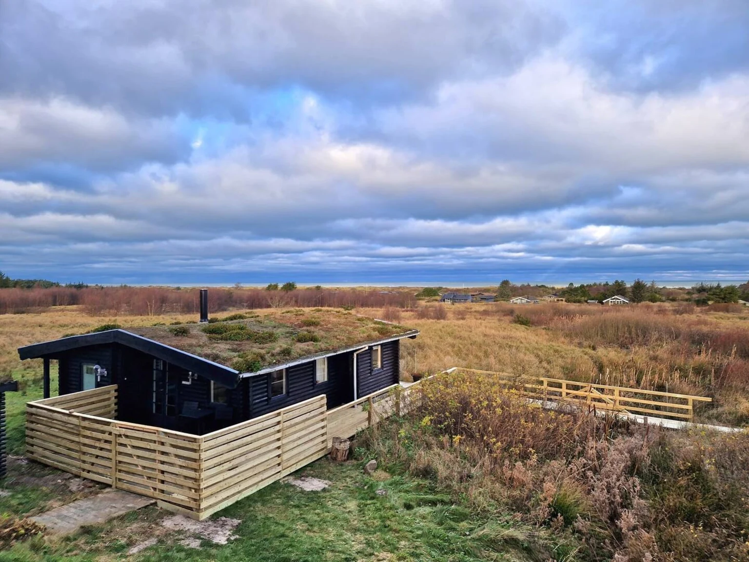 Luftbild: das Blockhaus mit Gründach und hellem Holzzaun in der Heide unter weitem Himmel