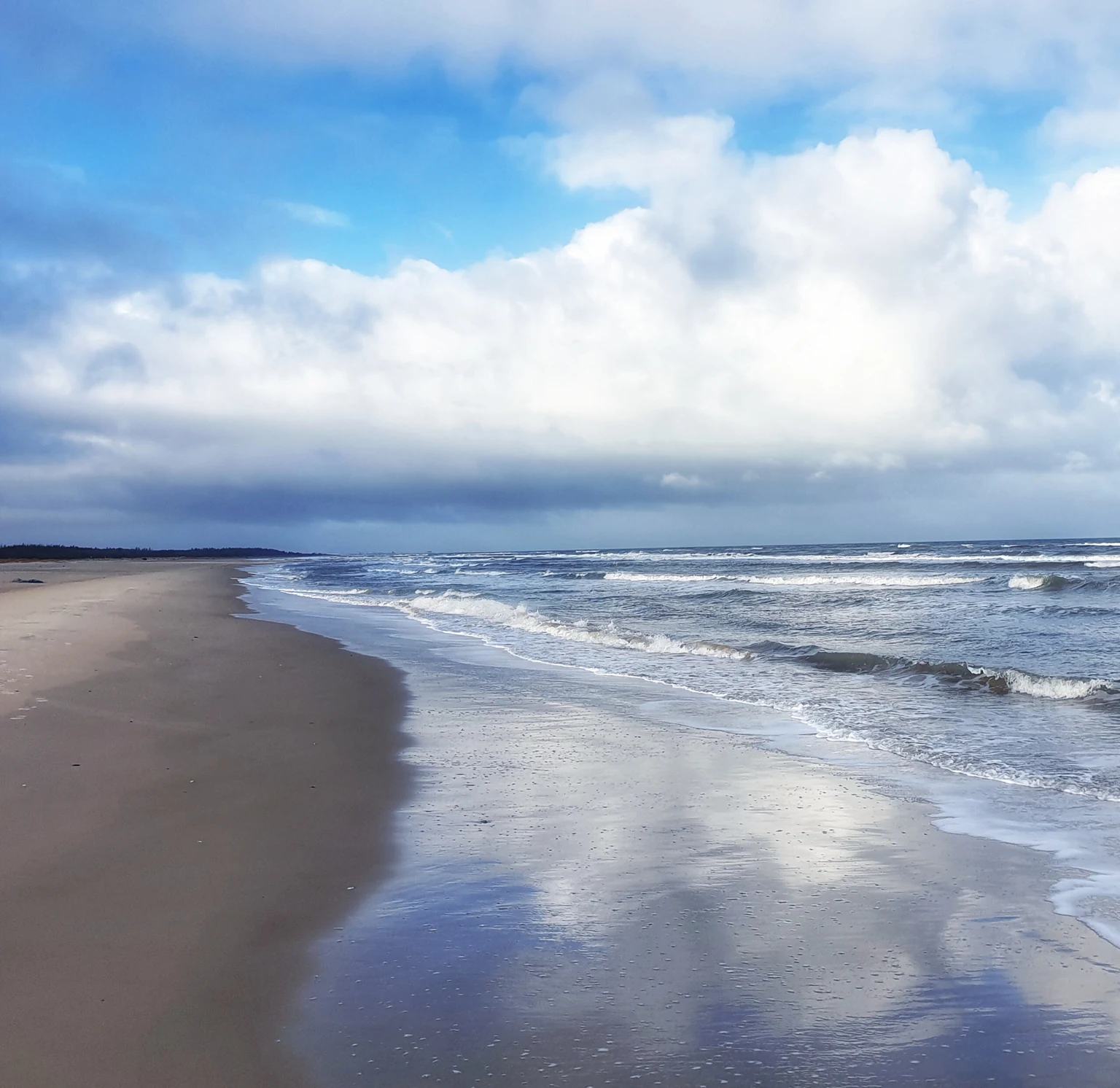 Sandstrand mit Wellen, Wolken spiegeln sich im nassen Sand