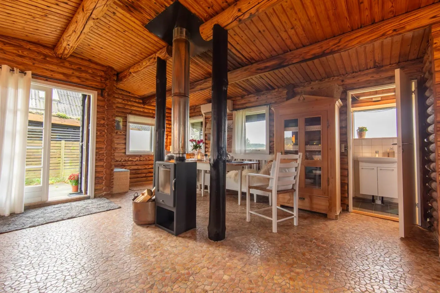 Wohnraum aus Rundholz mit Holzofen in der Mitte, Essplatz und Tür nach draußen